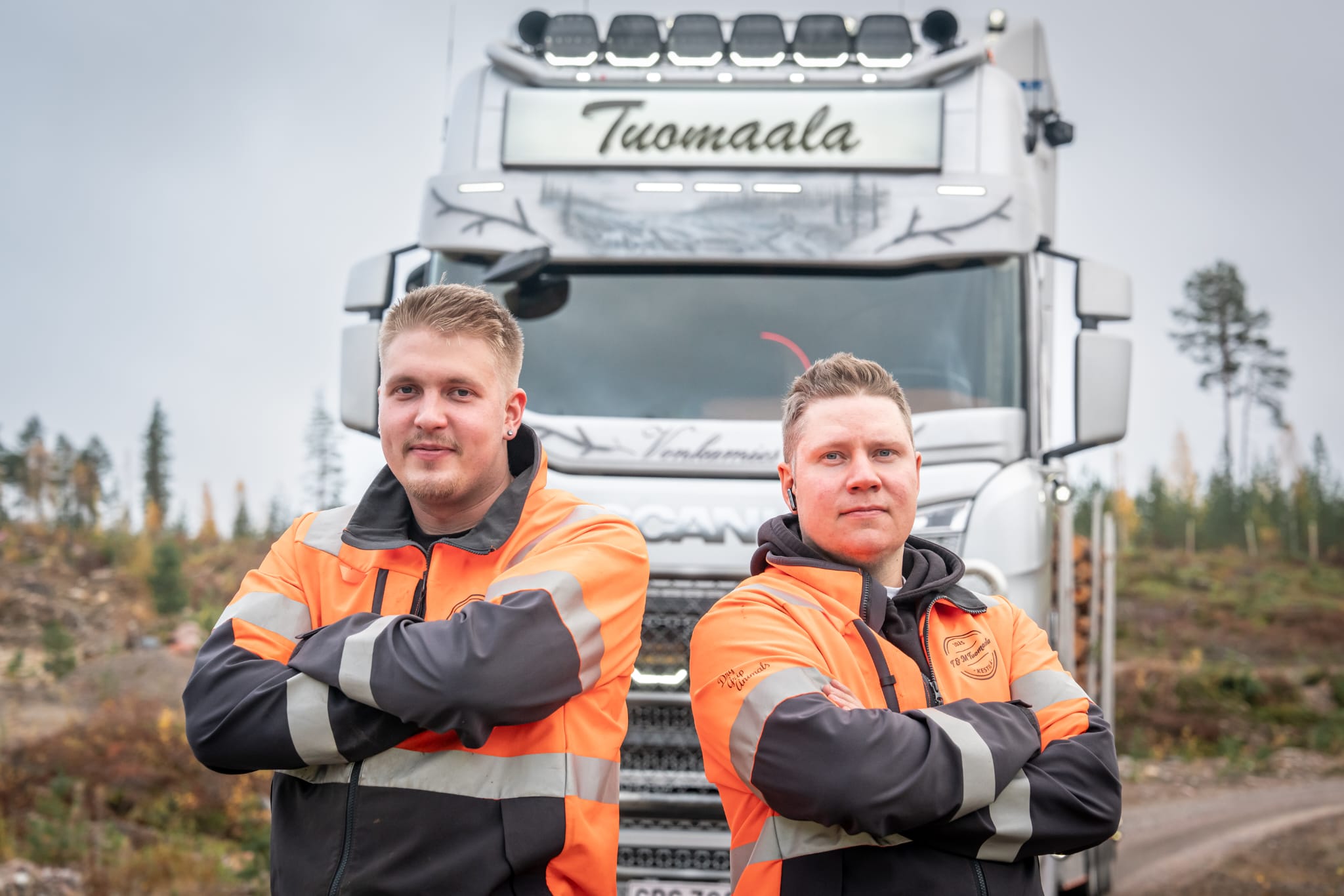 Kaksi Tuomaalan kuljettajaa huomiovaatteissa yrityksen Scania-puutavara-auton edessä.